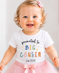 A smiling baby girl wearing a white t-shirt with text and a pink bow.
