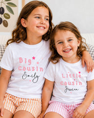 Two young girls wearing matching white t-shirts with personalized text, smiling and sitting together.