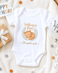 A white onesie with a cat design and the text "Halfway to One - Shepherd" printed on it, along with a "Happy Birthday" sign and confetti.