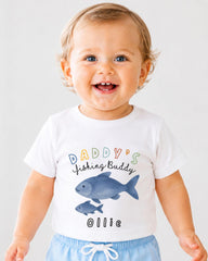 A smiling baby boy wearing a white t-shirt with a fish graphic and text that says "DADDY'S Fishing Buddy Ollie".