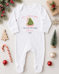 A white onesie with a Christmas tree design and text, surrounded by Christmas decorations and ornaments.