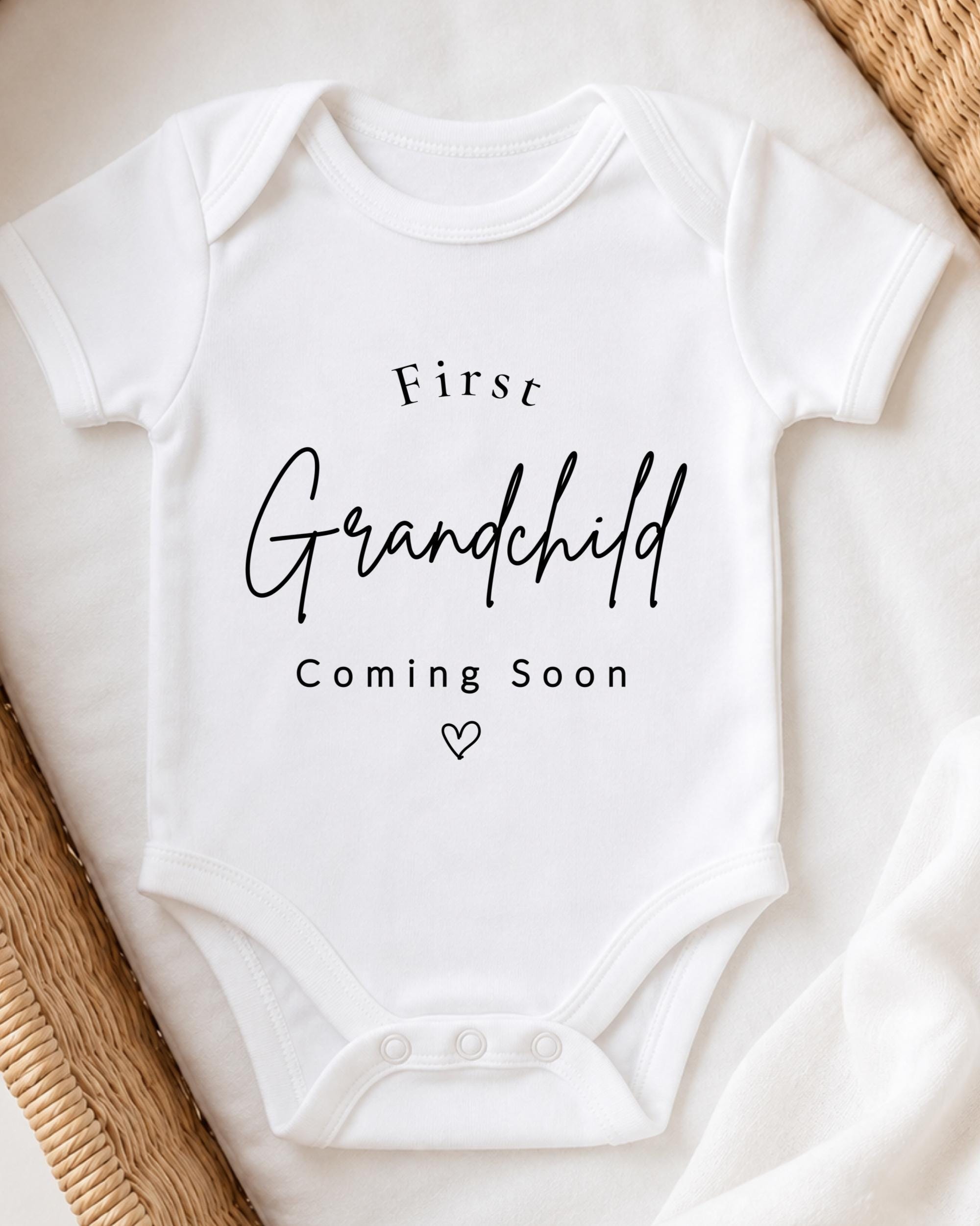A white onesie with the text "First Grandchild Coming Soon" printed on it, placed on a bed with a woven blanket.