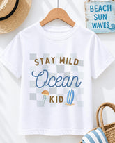 A white t-shirt with a graphic design that says "STAY WILD OCEAN KID" and includes a jellyfish and a surfboard. The t-shirt is hanging on a wooden hanger, and there is a straw hat and a beach sign in the background.