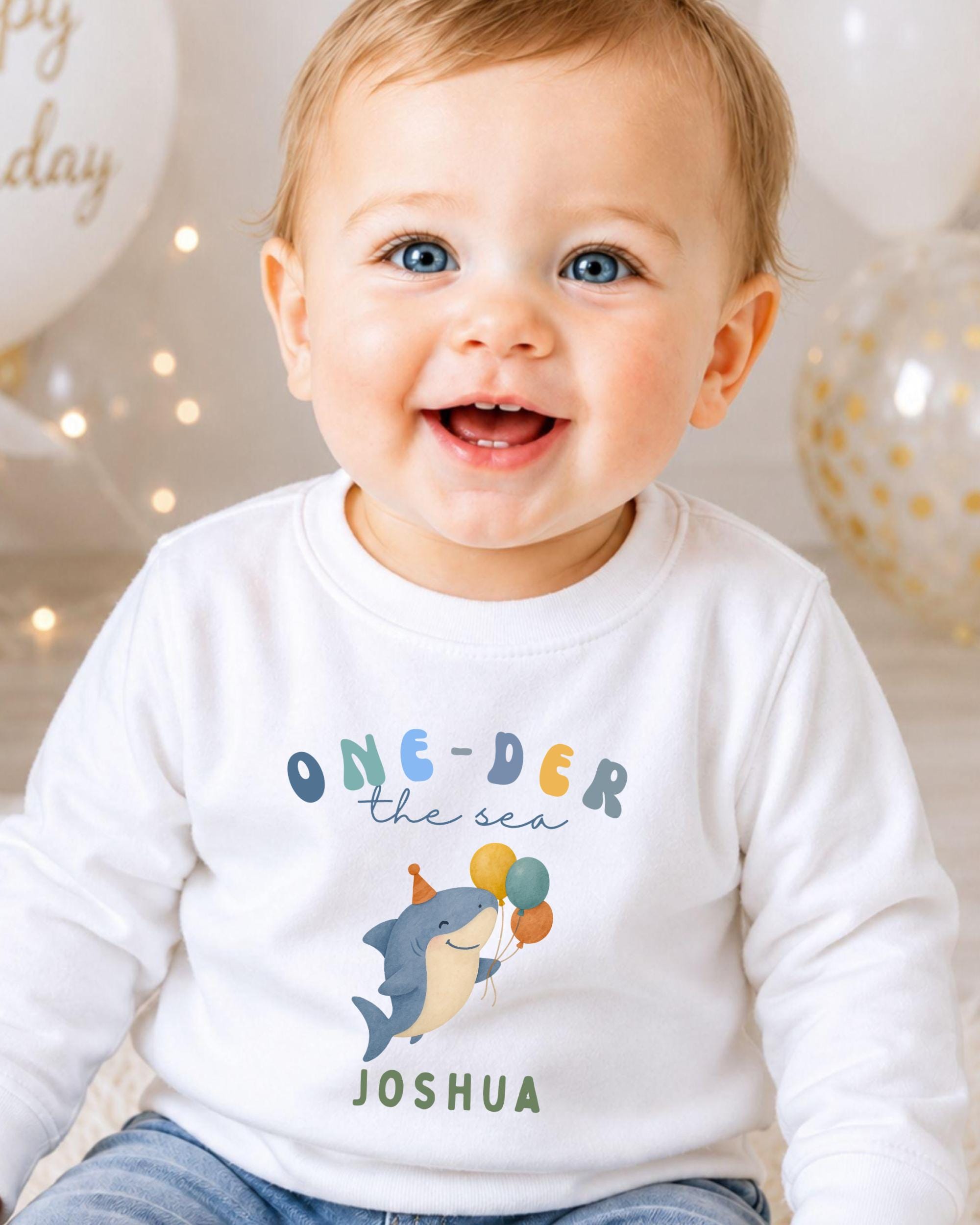 A smiling baby boy wearing a white shirt with a cartoon shark and the text "One-der the sea Joshua" printed on it.