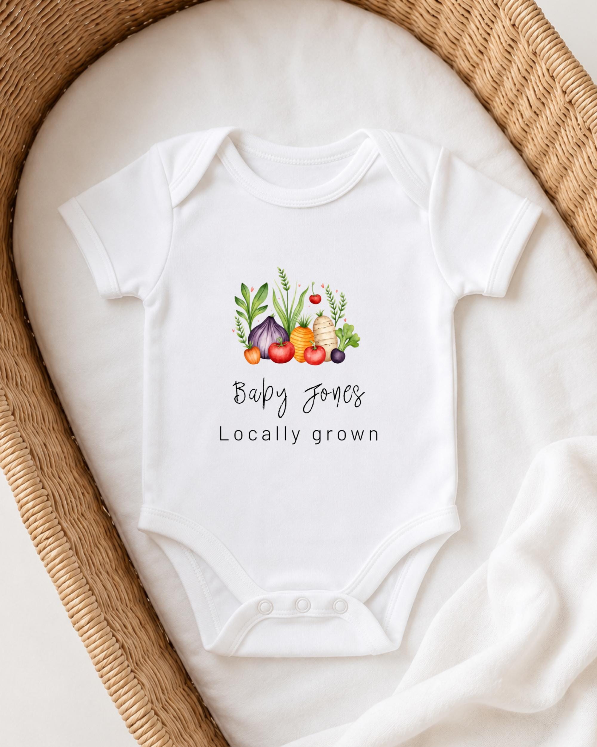 A white baby onesie with a graphic of vegetables and the text "Baby Jones Locally grown" printed on it, placed in a woven basket.
