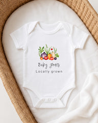 A white baby onesie with a graphic of vegetables and the text "Baby Jones Locally grown" printed on it, placed in a woven basket.