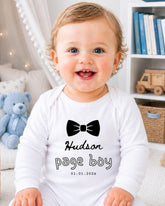 A smiling baby boy wearing a white onesie with a black bow tie and the text "Hudson page boy 01.01.2023" printed on it.