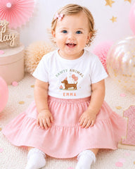 A smiling baby girl wearing a white t-shirt with a paw print design and a pink skirt, sitting on a pink and white background with balloons and confetti.