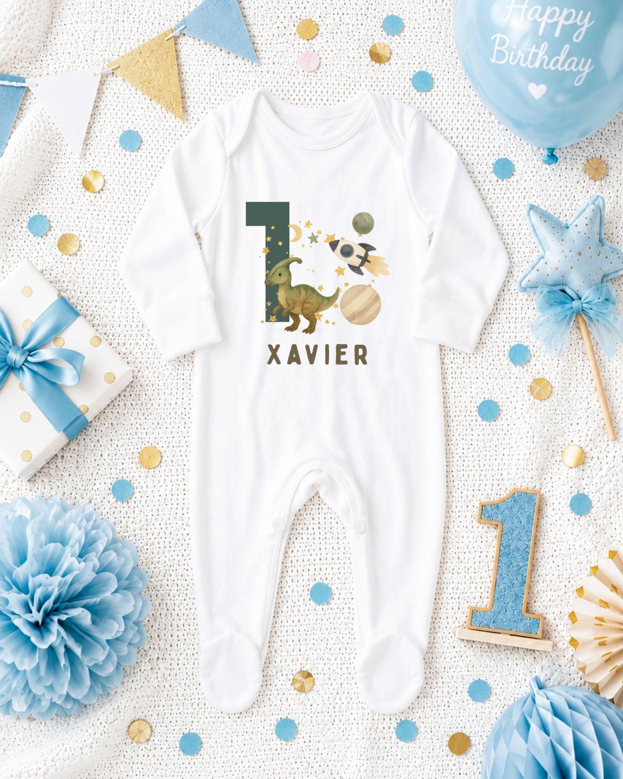 A white onesie with the number 1 and dinosaur designs, surrounded by birthday decorations such as a blue balloon, blue and gold confetti, and a blue and gold star.