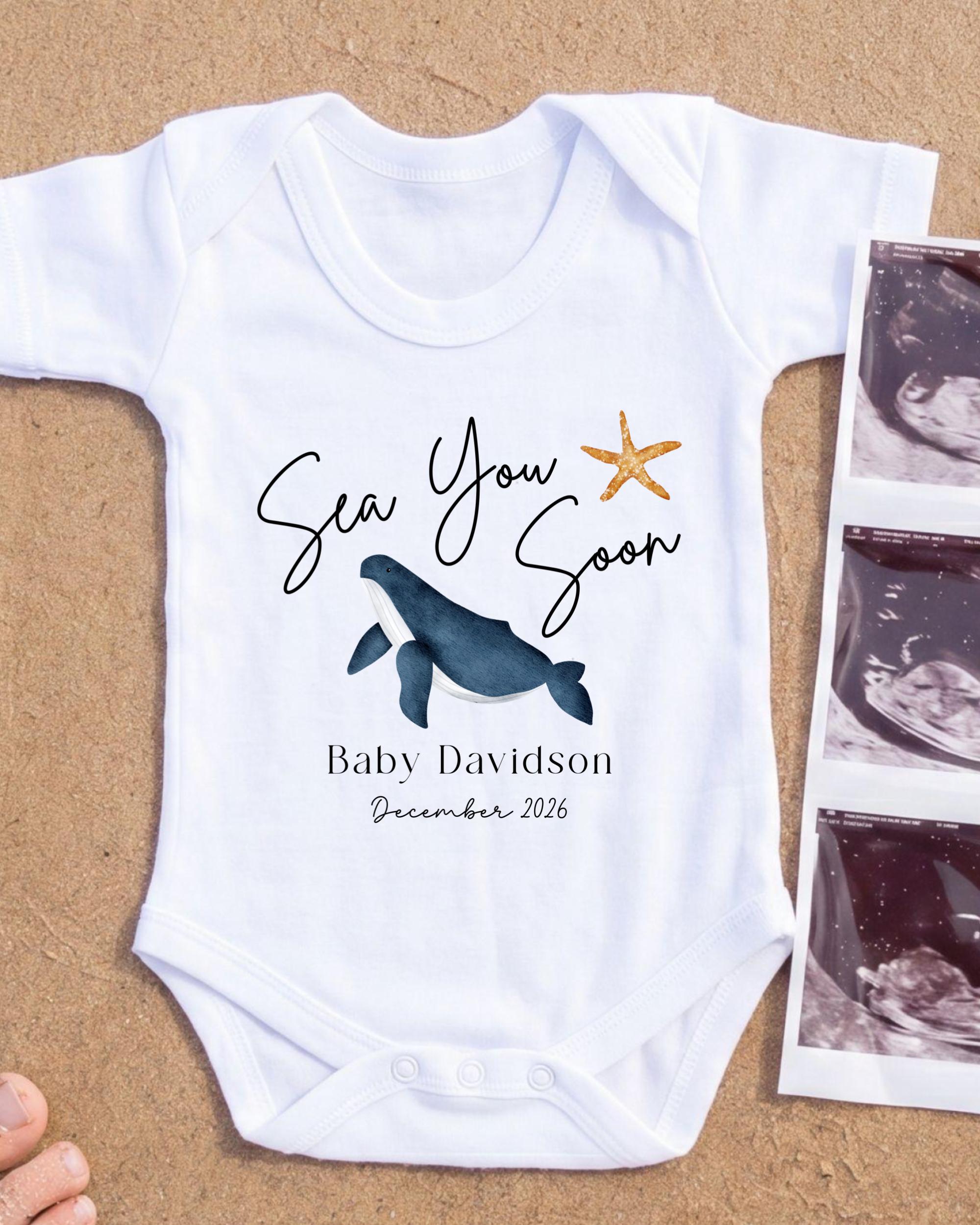 A white baby onesie with a graphic of a whale and a starfish, along with the text "Sea You Soon" and "Baby Davidson December 2026".