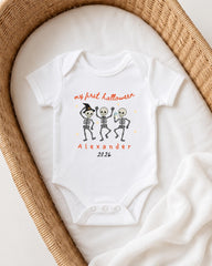 A white onesie with a Halloween-themed design, featuring three cartoon skeletons dancing and the text "my first Halloween Alexander 2026".