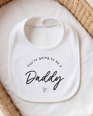A white bib with the text "You're going to be a Daddy" printed on it, placed on a white cloth.