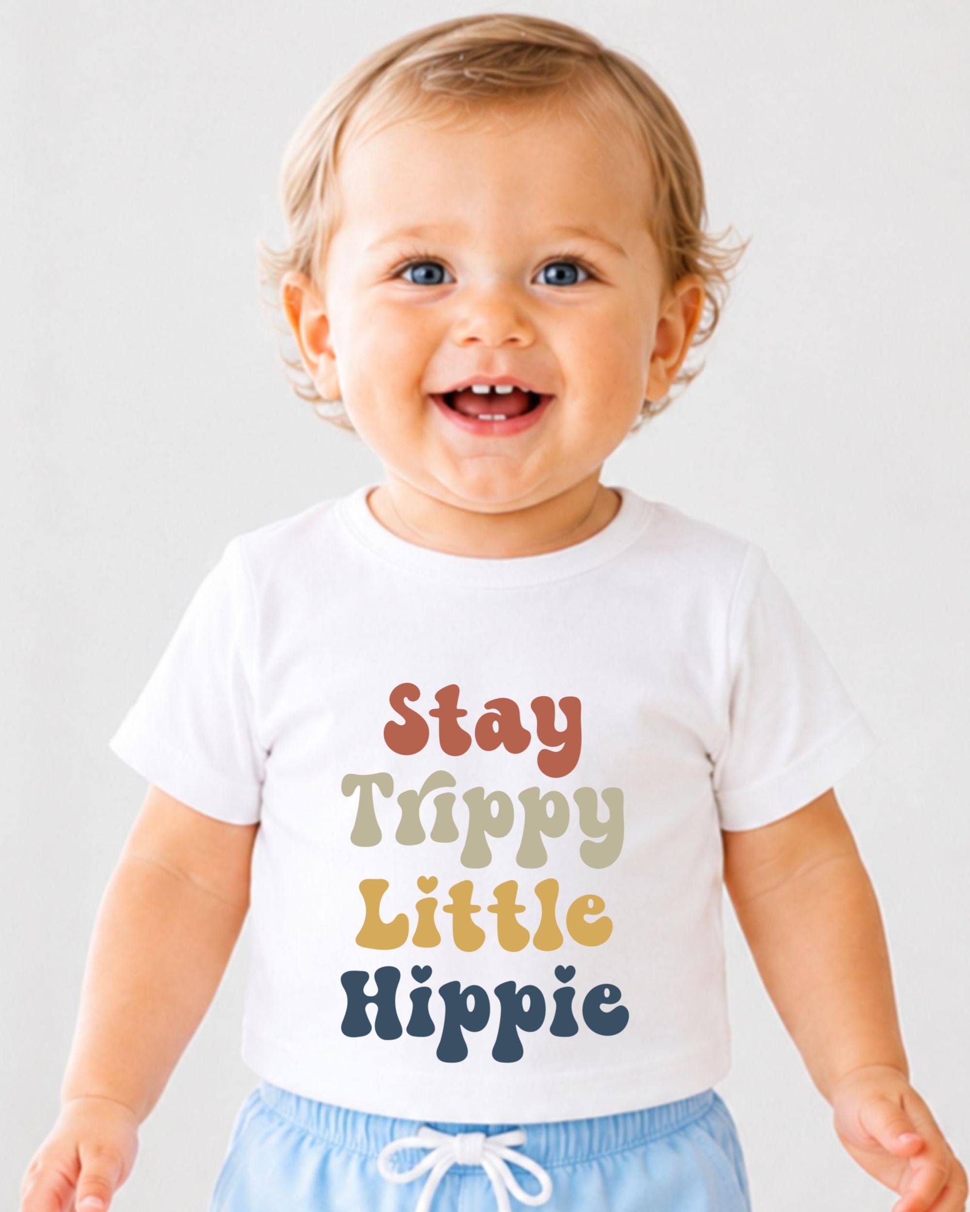 A smiling baby wearing a white t-shirt with the text "Stay Trippy Little Hippie" printed on it.