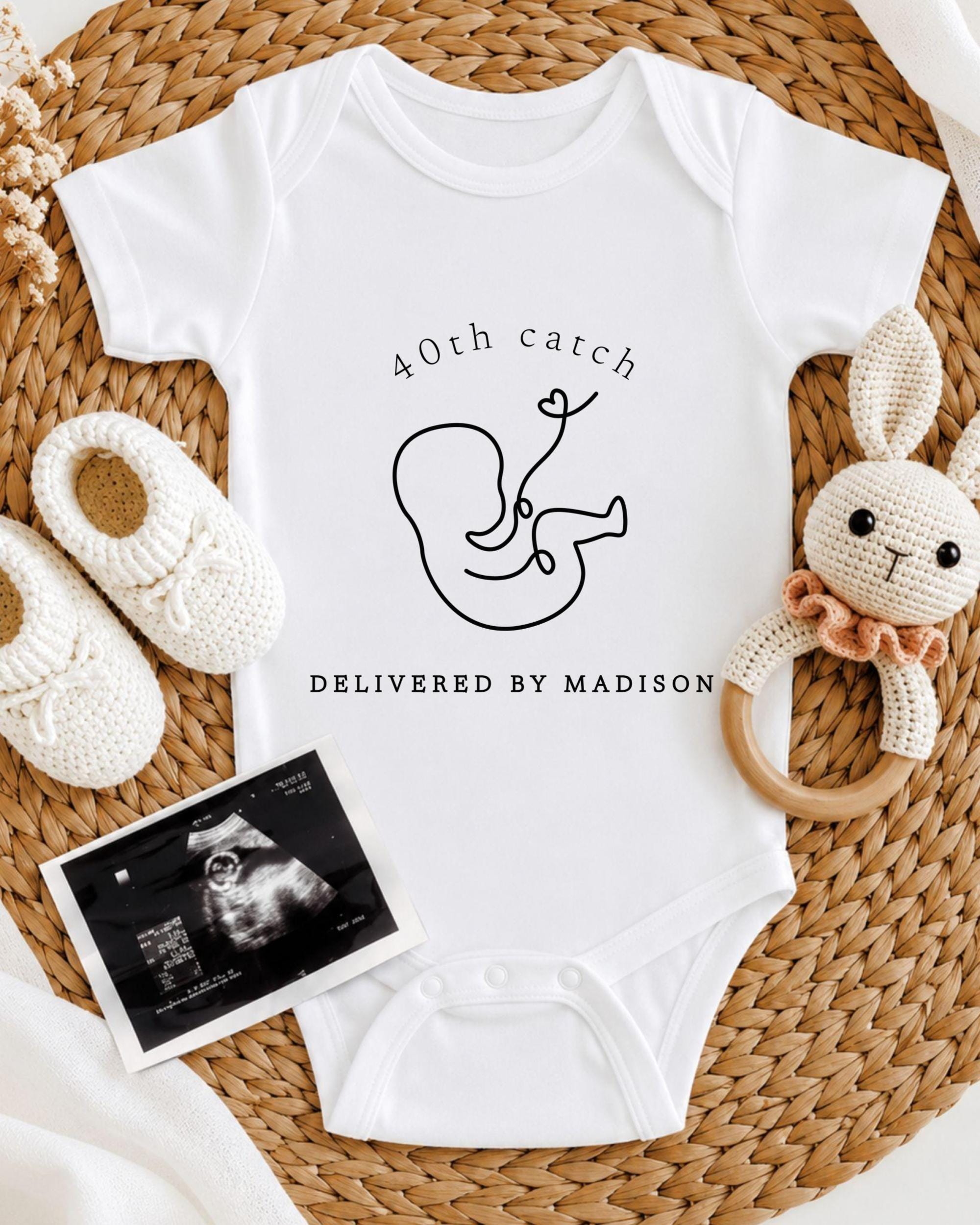A white onesie with a graphic of a baby and the text "40th catch DELIVERED BY MADISON" is displayed on a woven placemat, along with a crocheted rabbit toy and a black and white photograph of an Ultrasound image.