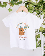 A white t-shirt with a cartoon image of a teddy bear holding a party hat and a balloon, along with the text "Half way to one" and "Liam 2026".