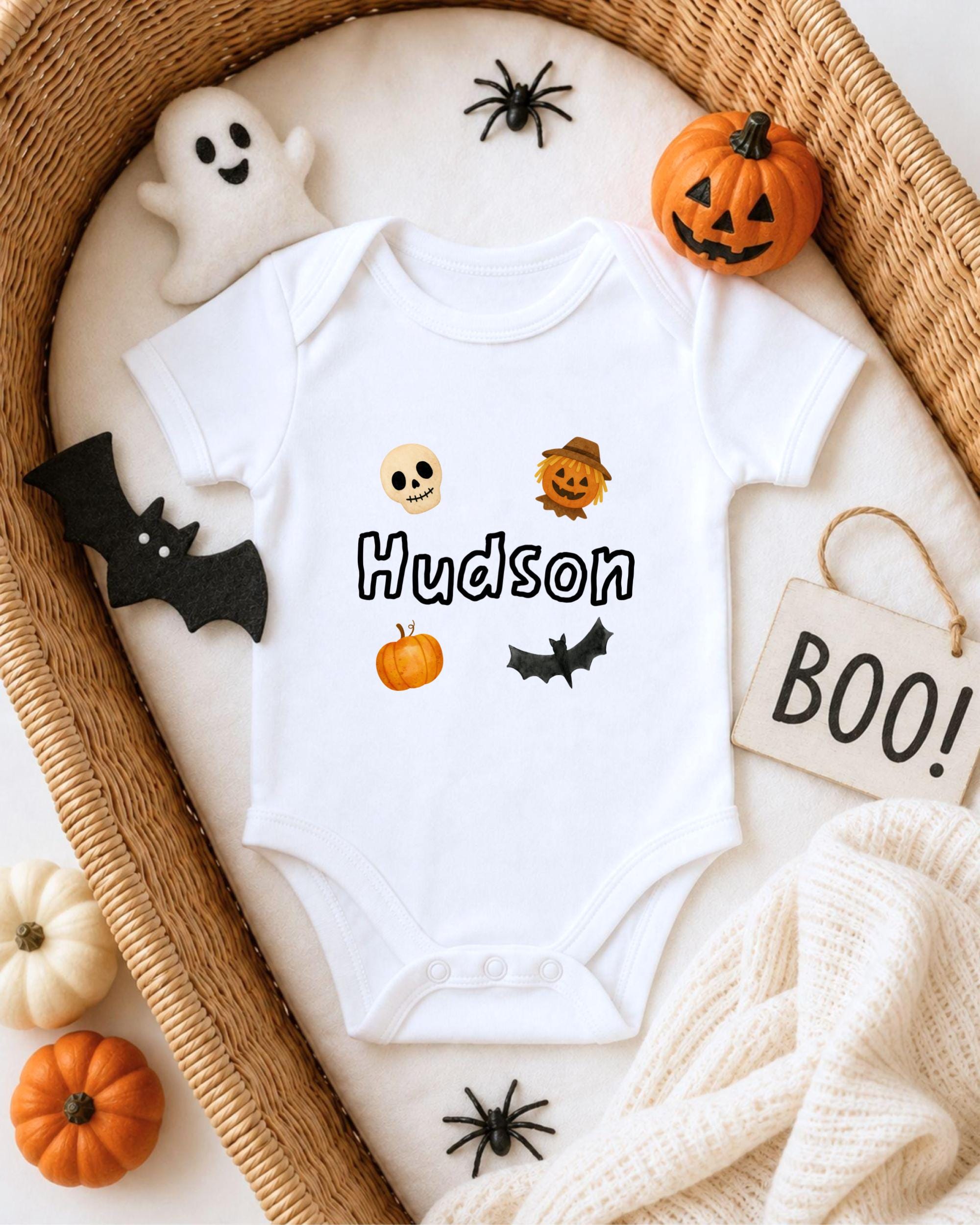 A white onesie with the name "Hudson" printed on it, surrounded by Halloween-themed decorations such as a ghost, a pumpkin, a bat, and a sign that says "BOO!".