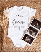 A white baby onesie with the text "BABY Harrison Coming Soon" printed on it, along with a black and white image of an ultrasound of a baby.