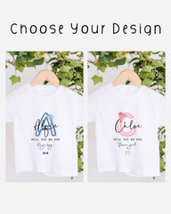 two white t-shirts hanging on a wooden hanger, each with a personalized message printed on them. The messages read "Will you be our Page boy" and "Will you be our Flower girl", suggesting that the t-shirts are intended as gifts or souvenirs for someone named Page boy and Flower girl.