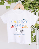 A white t-shirt with the text "HOLIDAY CREW Tenerife" printed on it, along with various summer-themed illustrations such as a sun, sunglasses, a beach ball, and a sandcastle.