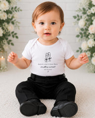 A smiling baby boy wearing a white shirt with a graphic and text, sitting on a white carpet in front of a floral background.