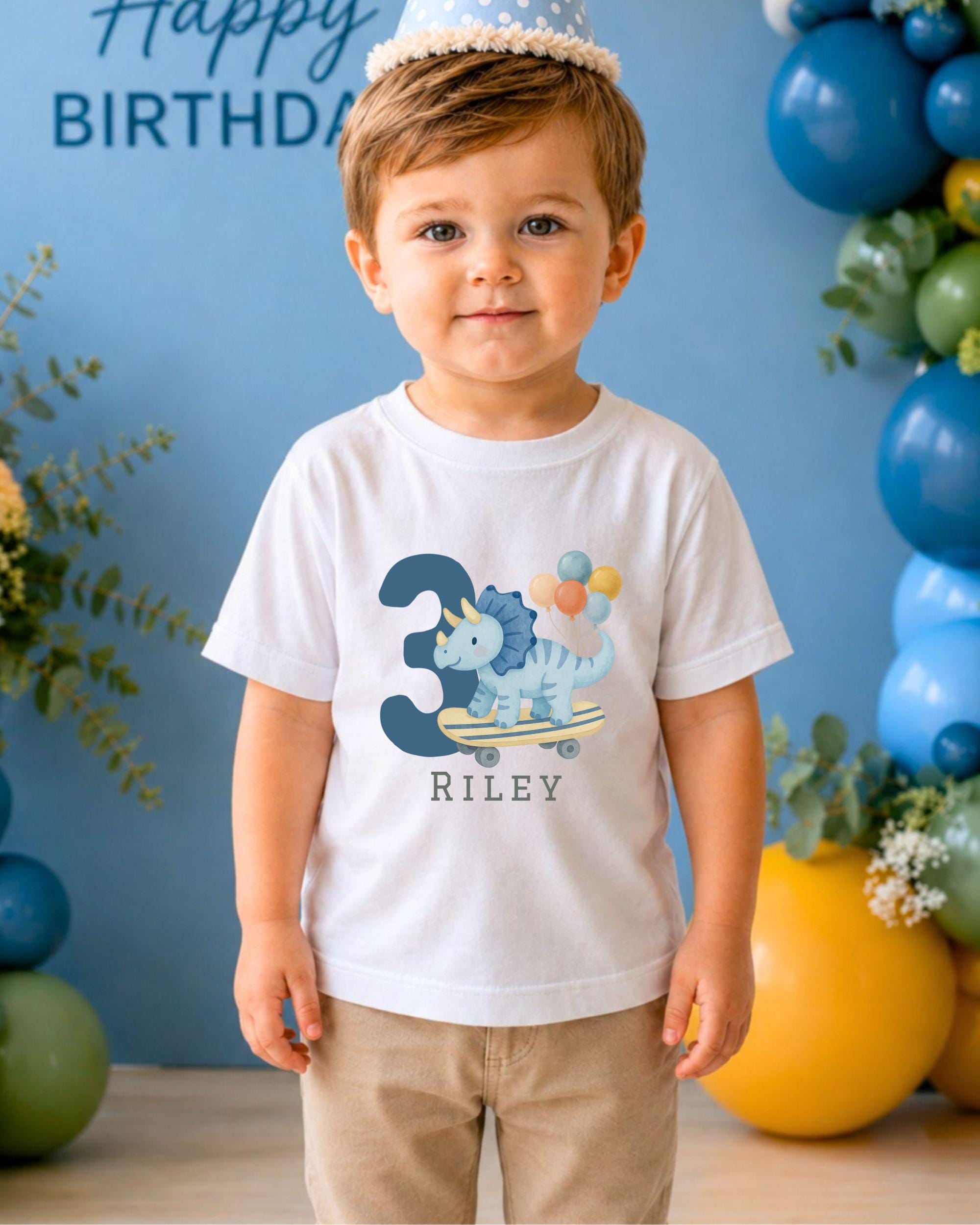 A young boy wearing a white t-shirt with a dinosaur graphic and the number 3, standing in front of a birthday party backdrop with balloons and greenery.