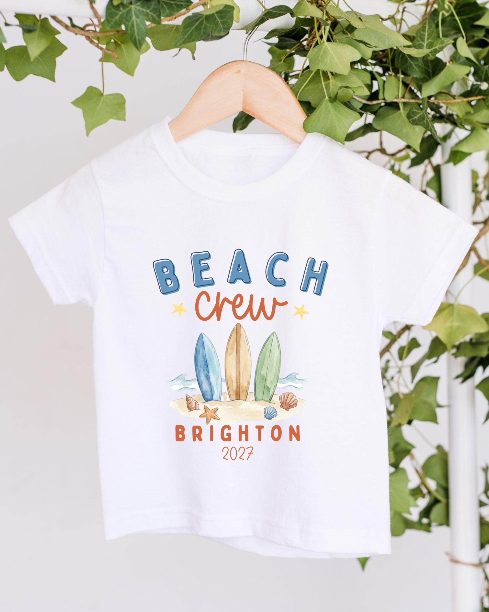 A white t-shirt with a graphic design featuring surfboards and beach elements, along with the text "BEACH CREW BRIGHTON 2027".