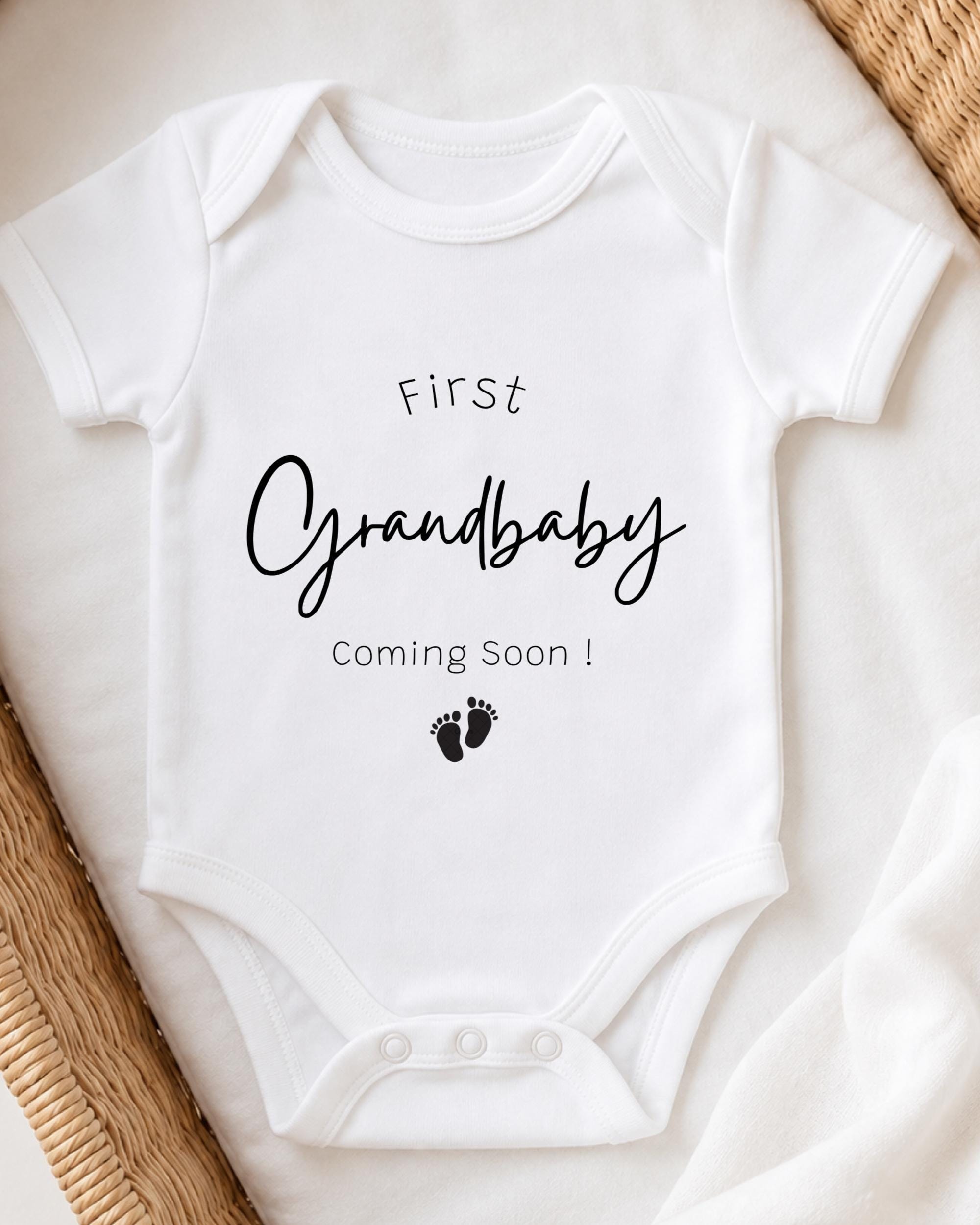 A white onesie with the text "First Grandbaby Coming Soon!" printed on it, along with a small footprint icon.