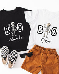A black t-shirt with a skeleton design and the name "Alexander" printed on it, a white t-shirt with the same skeleton design and the name "Oscar" printed on it, a pair of brown shorts, and a pair of black and white checkered shoes.