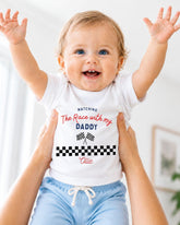 A smiling baby wearing a white t-shirt with a racing theme and text, being held up by someone's arms.