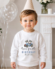A young boy wearing a white sweatshirt with a birthday hat and a car design, smiling at the camera.