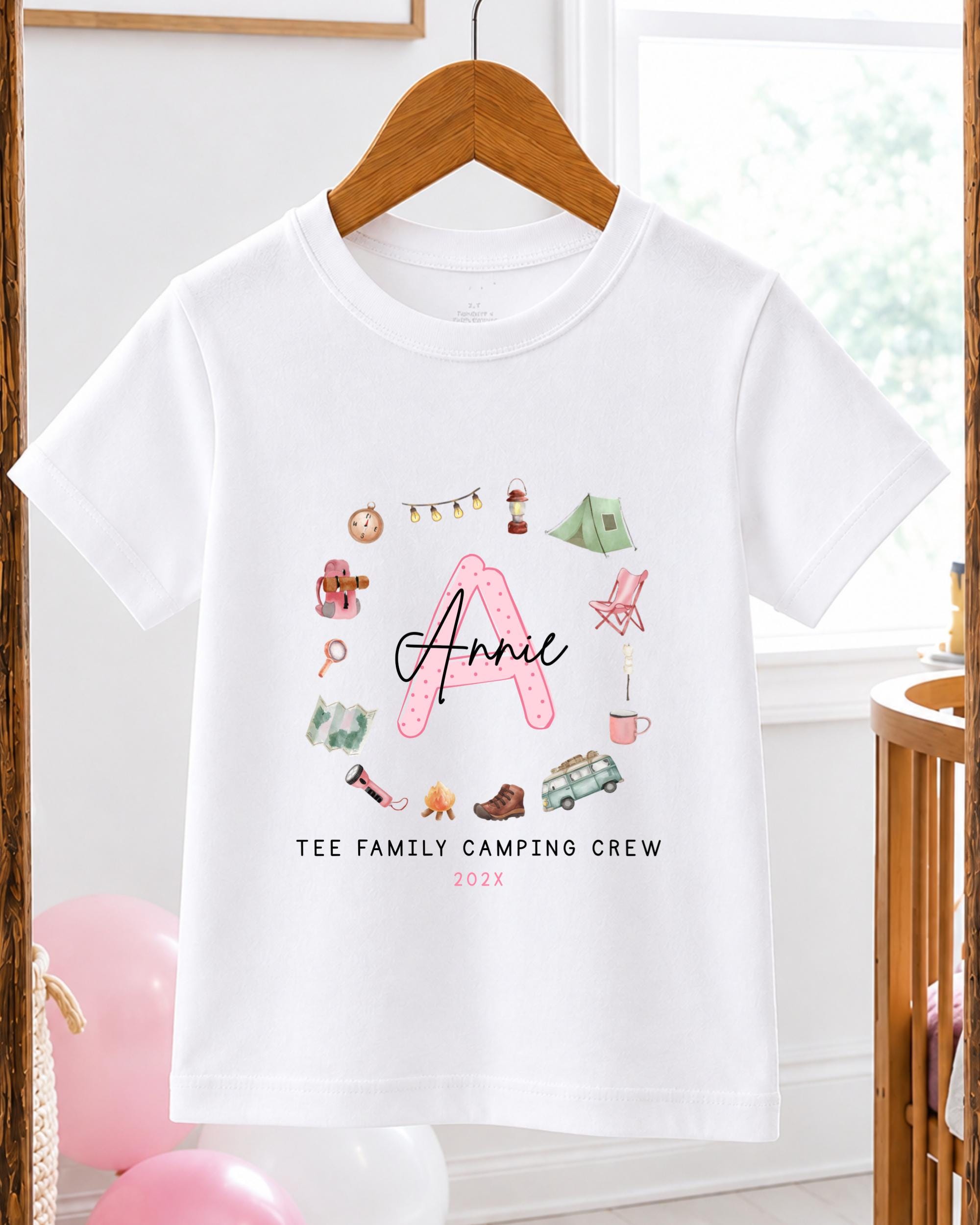 A white t-shirt with a personalized design featuring a pink "A" surrounded by camping-related items such as a tent, camping chair, and camping stove. The text on the t-shirt reads "A Annie Tee Family Camping Crew 2020".