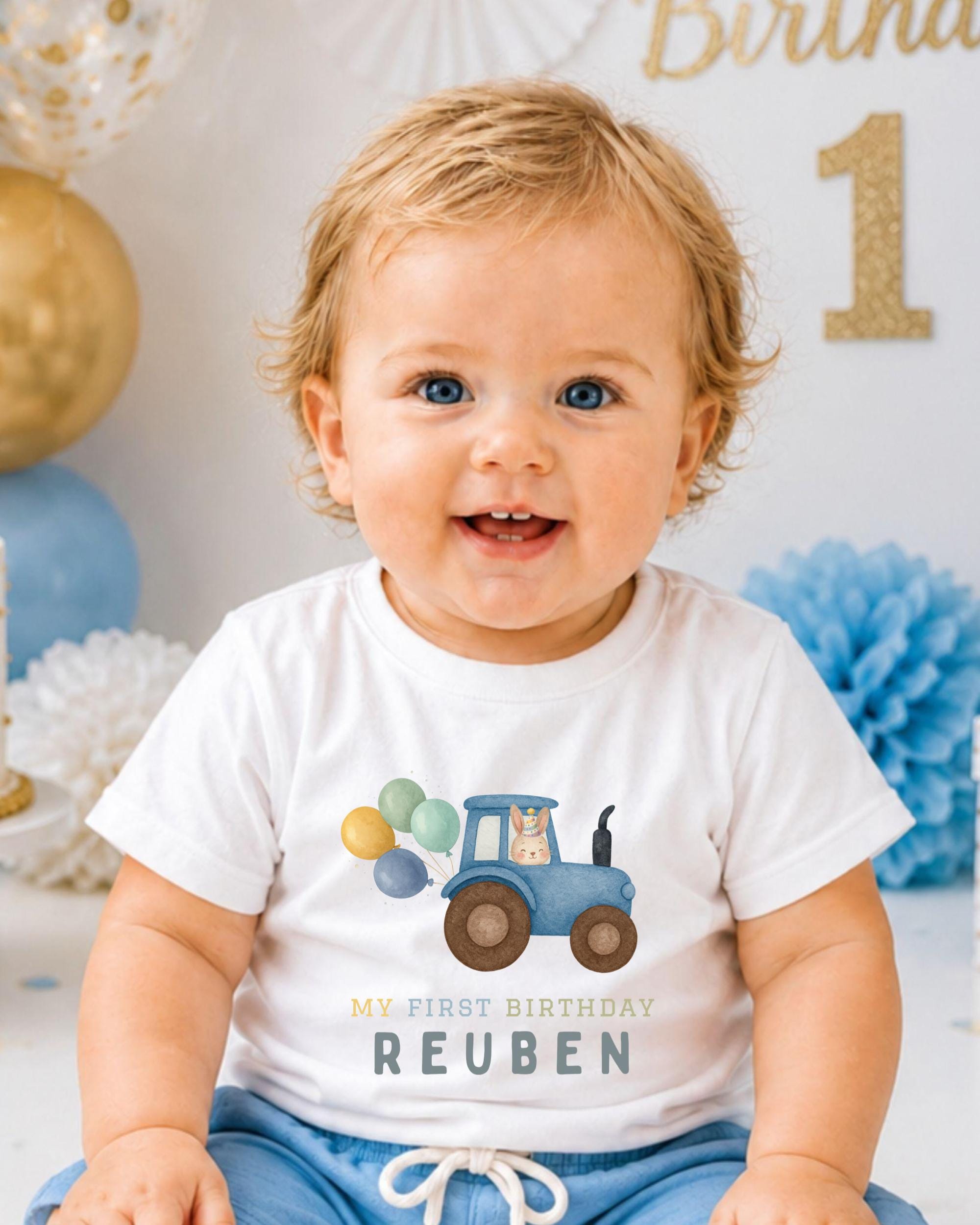 A smiling baby boy wearing a white t-shirt with a tractor graphic and the text "My first birthday Reuben" printed on it.