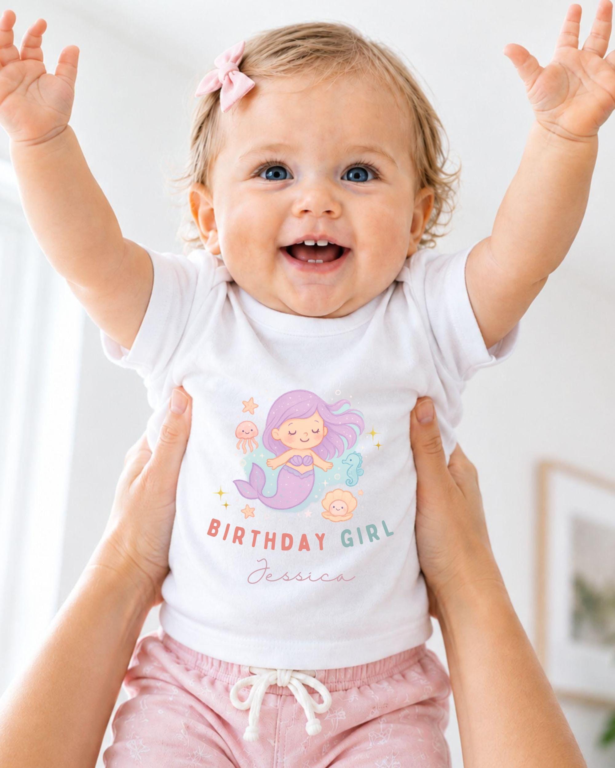 A smiling baby girl wearing a white t-shirt with a mermaid graphic and the text "Birthday Girl Jessica" is being held up by someone's arms.
