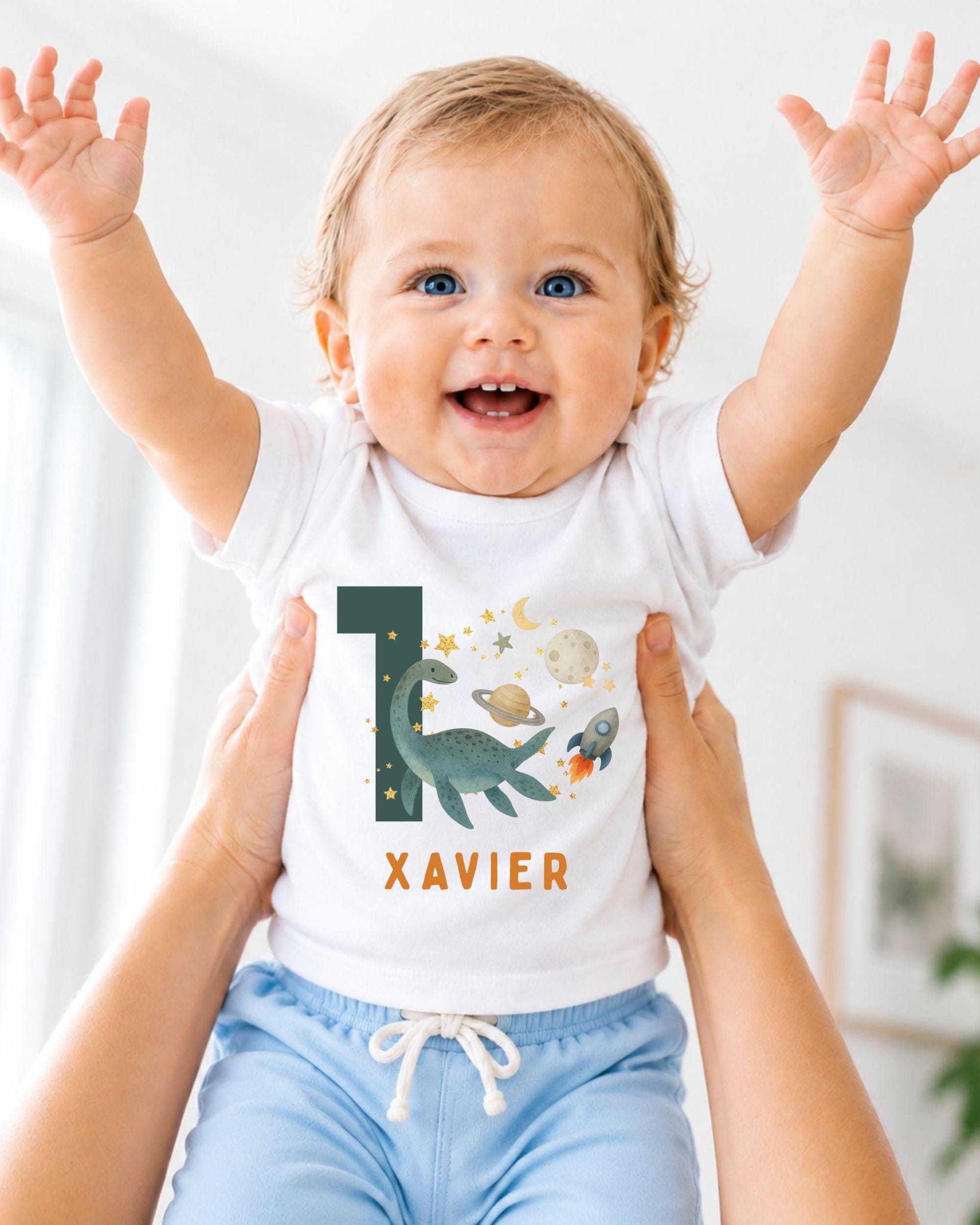 A smiling baby wearing a white t-shirt with a dinosaur and space-themed design, being held up by someone's arms.