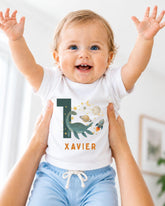 A smiling baby wearing a white t-shirt with a dinosaur and space-themed design, being held up by someone's arms.