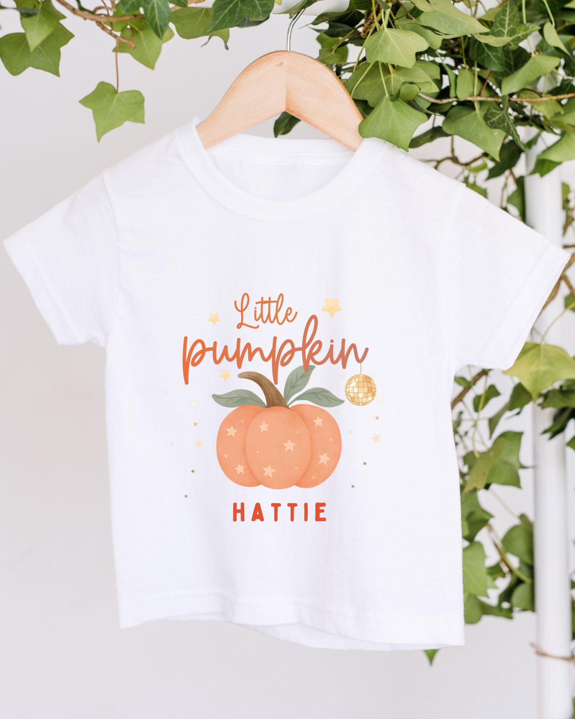A white t-shirt with a pumpkin graphic and text that says "Little pumpkin" and "HATTIE" printed on it.