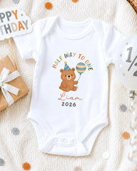 A white onesie with a cartoon bear and a balloon, along with a "Happy Birthday" sign and confetti, suggesting a celebration of a child's first birthday.