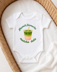 A white onesie with a graphic of a green drink and the text "Mama's favourite matcha date" printed on it, placed on a white blanket.