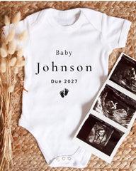 A white baby onesie with the text "Baby Johnson Due 2027" printed on it, along with a black and white image of an ultrasound scan.