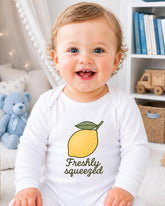 A smiling baby wearing a white onesie with a lemon graphic and the text "Freshly squeezed" is sitting in a room with bookshelves and a teddy bear.