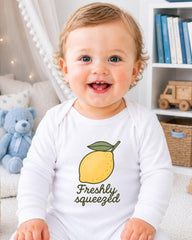 A smiling baby wearing a white onesie with a lemon graphic and the text "Freshly squeezed" is sitting in a room with bookshelves and a teddy bear.