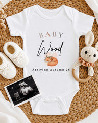 a baby onesie with the text "BABY Wood Arriving Autumn 26" printed on it, along with a crocheted rabbit toy and a baby bootie. There is also an Ultrasound image of a baby's belly.