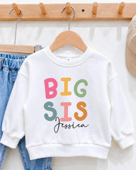 A white sweatshirt with the text "BIG SIS Jessica" printed on it, hanging on a wooden hanger.
