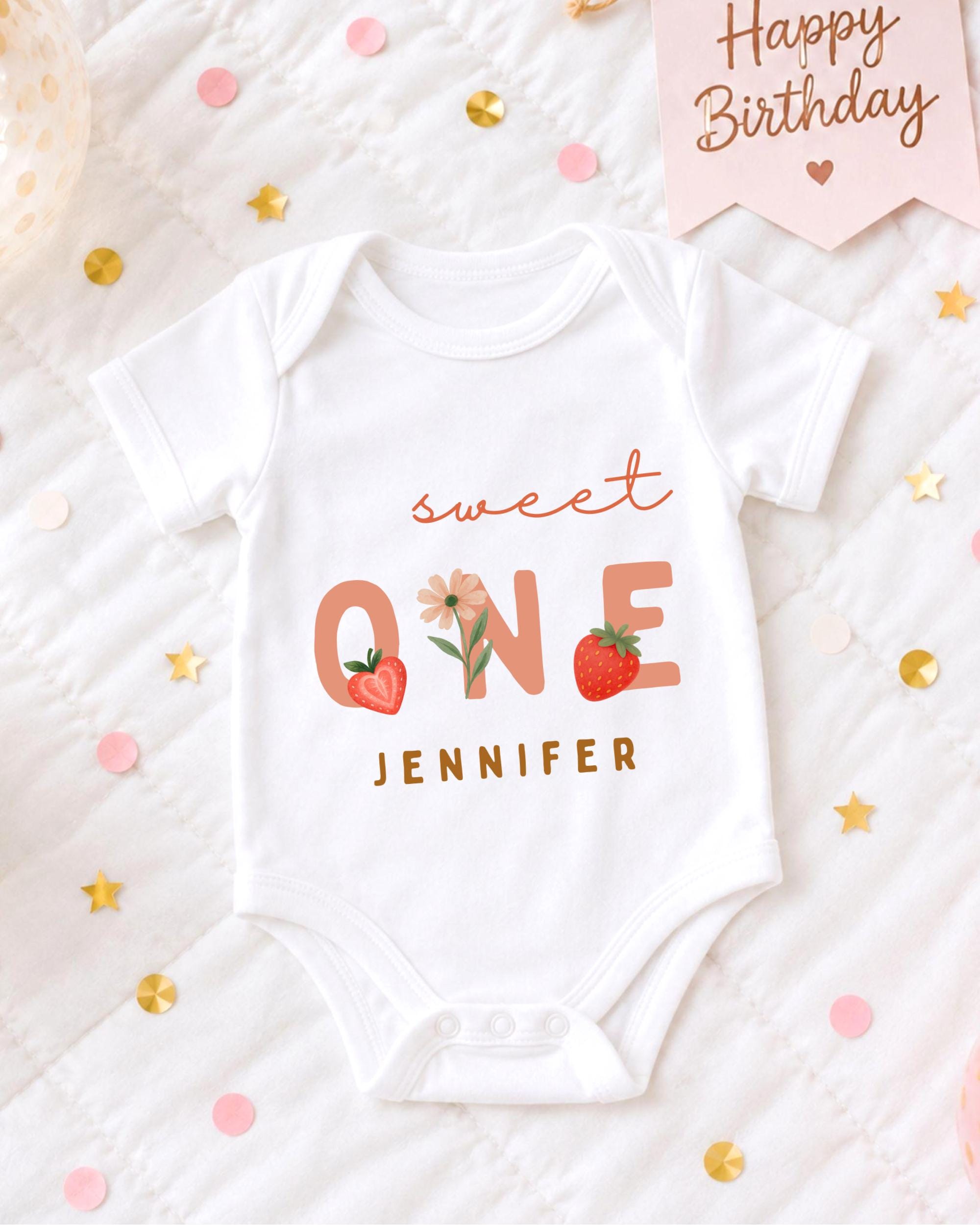 A white onesie with the text "Sweet One" printed on it, surrounded by confetti and a "Happy Birthday" sign.