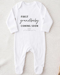 A white onesie with the text "FIRST GRANDBABY COMING SOON" printed on it.