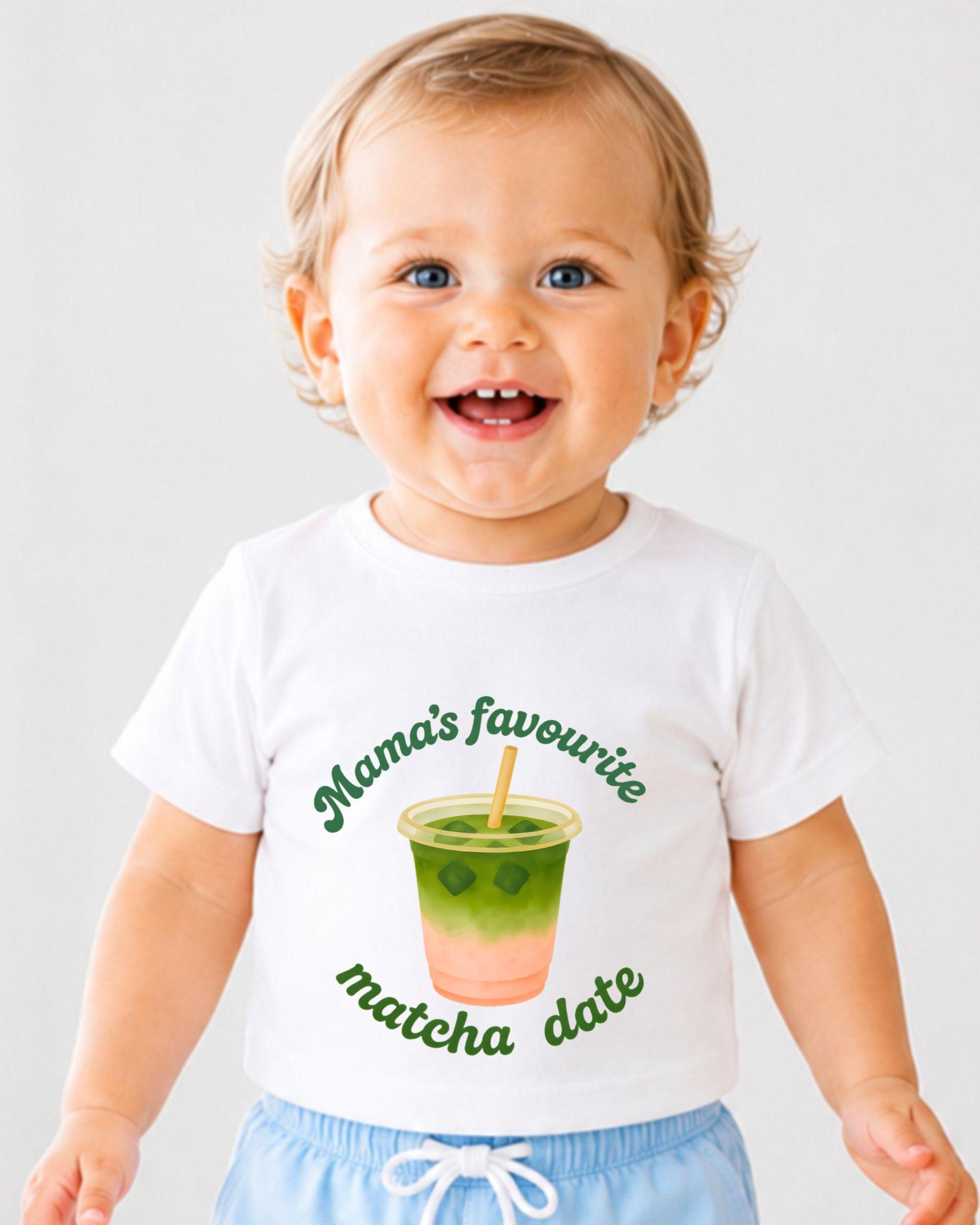 A smiling baby wearing a white t-shirt with a graphic of a green drink and the text "Mama's favourite matcha date" printed on it.