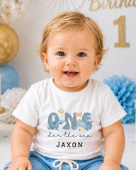 A smiling baby boy wearing a white t-shirt with a graphic design and text, sitting in front of a birthday party backdrop with balloons and a "1" sign.