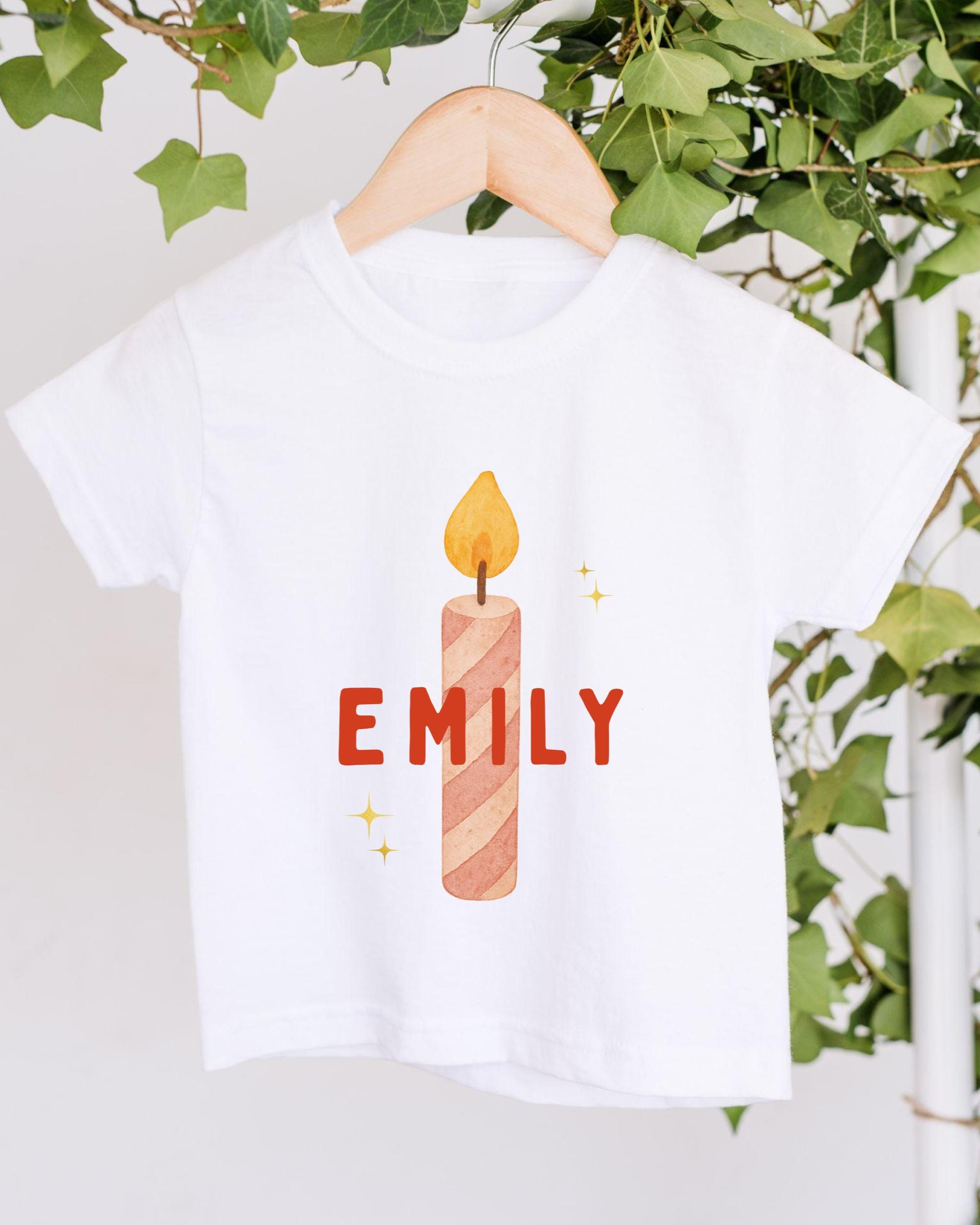 A white t-shirt with a graphic design of a lit candle and the name "EMILY" printed on it, hanging on a wooden hanger.