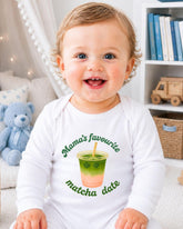 A smiling baby wearing a white onesie with a graphic of a green drink and the text "Mama's favourite matcha date" printed on it.