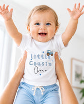 A smiling baby wearing a white shirt with the text "Little Cousin Oscar" is being held up by someone's arms.